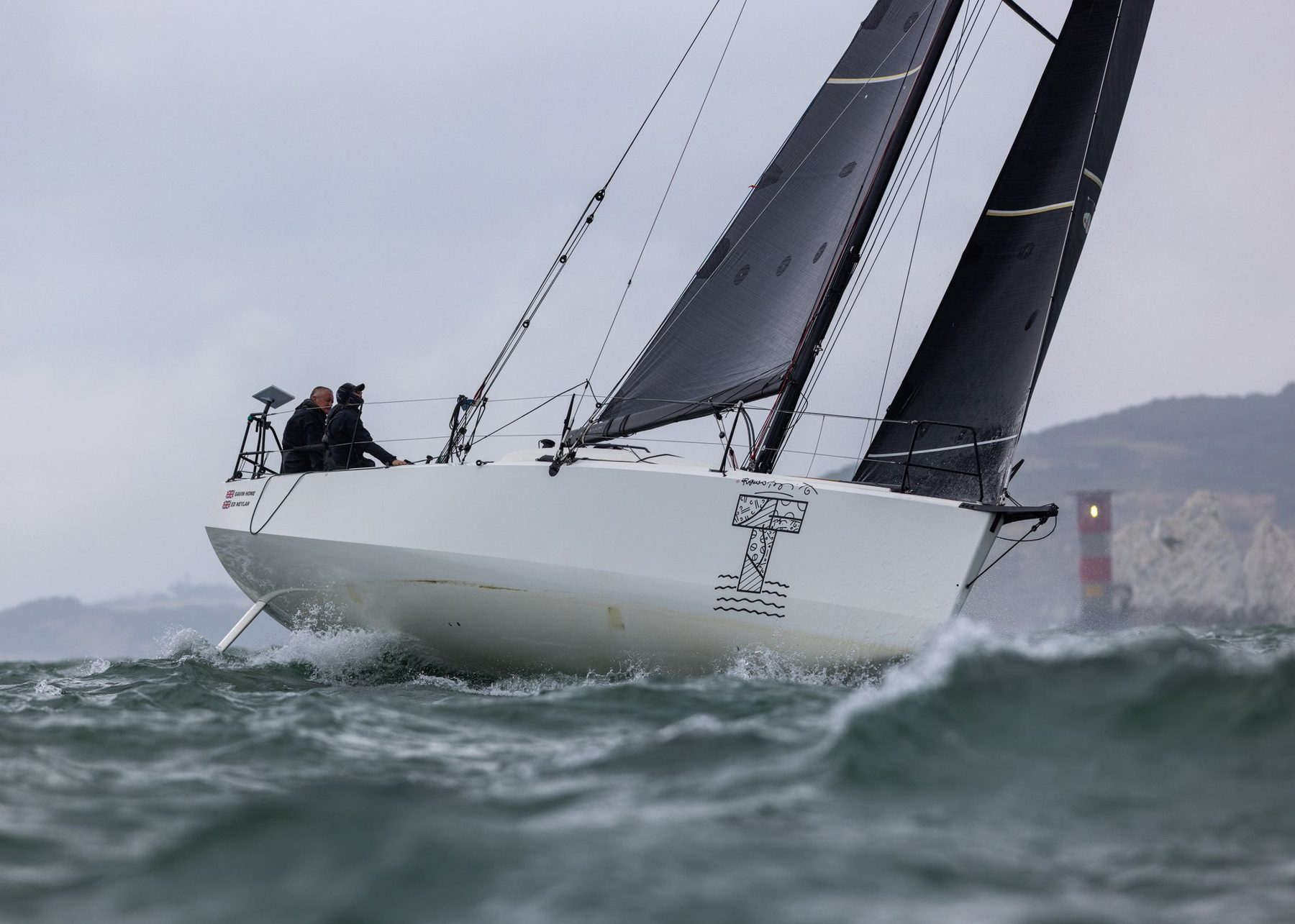A racing yacht heeled hard on a grey day, sailing past the Needles lighthouse at the western exit of the Solent